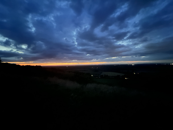 The end of Friday 26 July. The last light is a narrow orange band across the middle of the picture. Above, clouds largely fill a blue sky, looking inky blue as the day ends. Below, the darkness of the hillside and in the distance a few lights from houses on the lower ground below. 