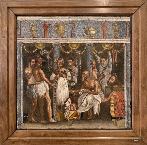 A mosaic showing a lavish party in a lavishly decorated room. Several men stand or sit listening to a flute player, masks can be seen around them, behind the group a man seems to don a costume with the help of another, seemingly actors getting ready to perform. 