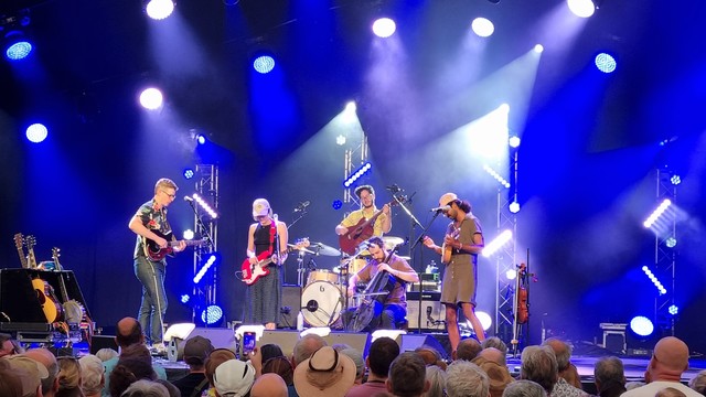 A band on stage, lit by glowing blue white beams from behind. Mandolin, cello, guitar, bass, electric guitar. 