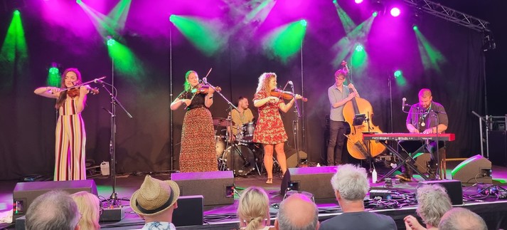 Three fiddlers on the left, a pianist and double bassist on the right, and drummer behind. All lit with green and purple lights.