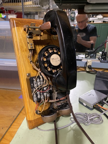 Close-up of a vintage rotary telephone mounted on a wooden base, with the internal wiring exposed. In the background, a person is working on something at a cluttered desk.