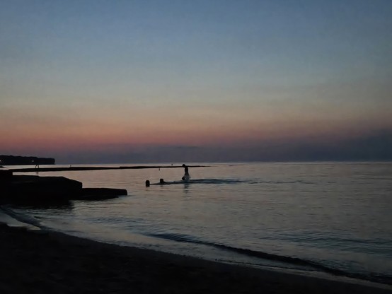 3 people swimming in the lake. The water is like glass. Like dark smokey glass. The Sun is below the horizon. The camera is pointed at WNW. The sky is dark at the horizon. Above the horizon, we get pastel oranges followed up with blue- grey