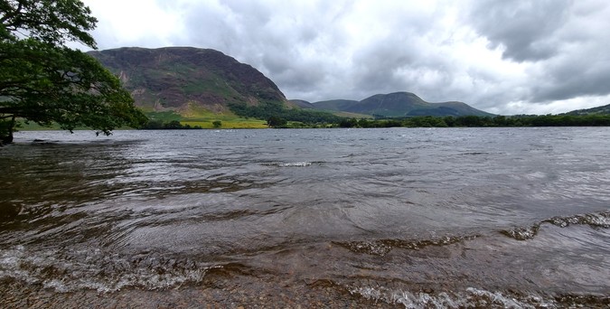 Crummock Water is choppy with a stiff breeze that crests the little waves in white, and blows grey clouds over the green crags on the other side of the lake. A pebble beach is just visible at the bottom of the frame, whilst a majestic oak tree hangs over the lake on the left.