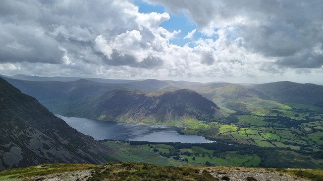 Crummock Water and Mellbreak. A view from high up of the lake with fells all around. Patches of sunlight pick out the small green fields, whilst the brown fells loom on the shore. Broken clouds are not yet threatening rain, but there is promise there.