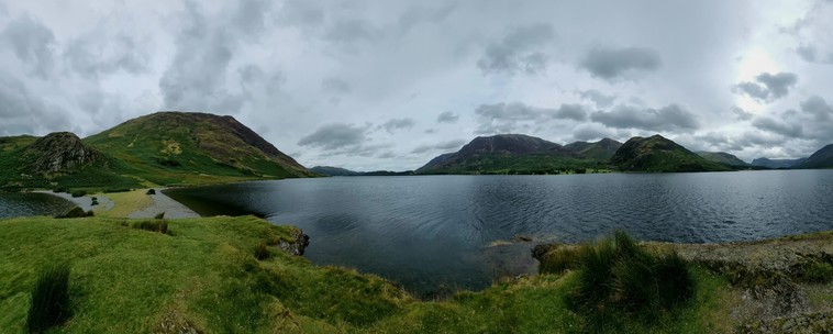 A panoramic view of a lake from close to the water. Grassy hummocks in the foreground lead to the shore via a gravel spot. Pale grey clouds are mirrored in the choppy lake surface. Beyond are bare green fells.