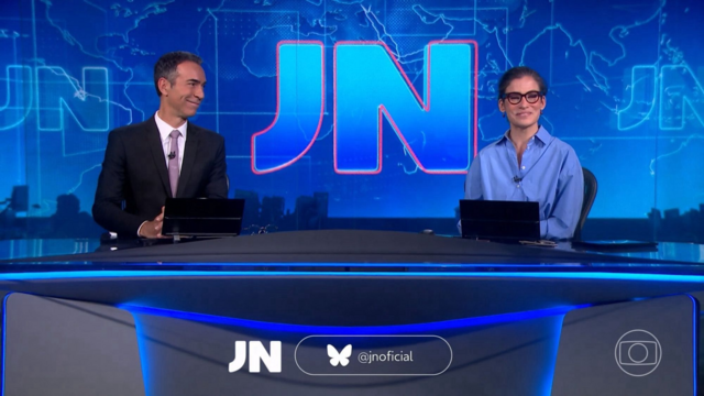 Two TV anchors (man and woman) on a TV news set all blue with a grez table, smiling. The Bluesky handle for the program appears on the screen