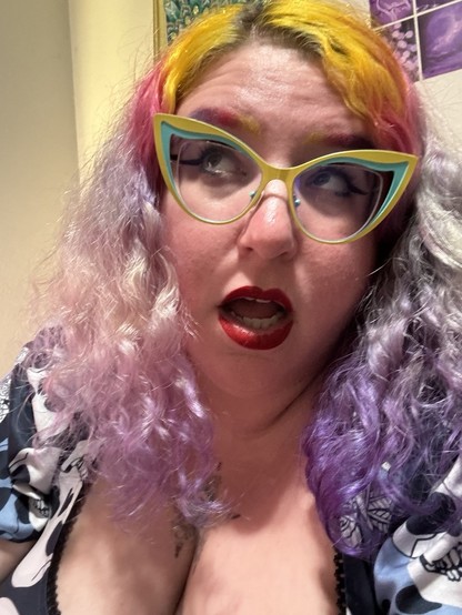 A picture of Stephanie looking up and to the side with her mouth open. She has long yellow, pink, and purple hair, matching eyebrows, long black winged liner under yellow and teal cat eye glasses, and red lips. She’s wearing a dark blue dress with white and light blue skulls on it.