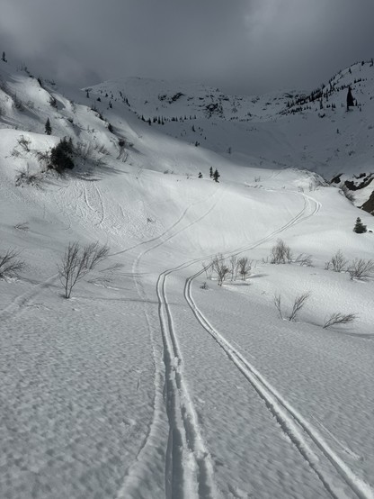 A big, open snowy mountain bowl. The foreground is lit by sun while the background is covered in cloud shadow.  two sets of ski tracks descend through the bowl toward the camera.