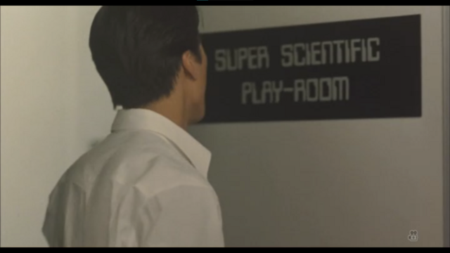 A person with black hair and a white shirt stands looking at a sign on a door, written in computer stencil lettering, white on a black background:

SUPER SCIENTIFIC
PLAY-ROOM