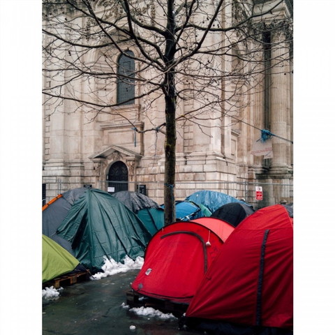 A photograph of colourful tents outside a cream stone building (St Paul's in London). Snow surrounds the edges of the tents. A tree stands in the centre of the image.