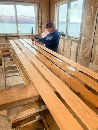 Interior of a cabin under construction. A stack of tongue and groove ceiling lumber sits on racks. An Inuk woman is applying urethane on the lumber with a paint brush. 