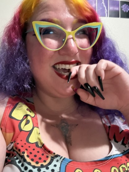 A picture of Stephanie with one finger between her teeth smiling and looking up and to the side. She has long wavy yellow, pink, and purple hair, matching eyebrows, long black winged eyeliner under yellow and teal cat eye glasses, and red lips. Her nails are long, black, and coffin shaped. She is wearing a red dress covered in comic book sound effects.