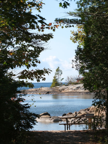 Approaching a lake through the forest. Trees left and right are offering just  a glimpse of the water
There is a picnic table on a sandy beach and a rocky tip of land jutting out into the bay.