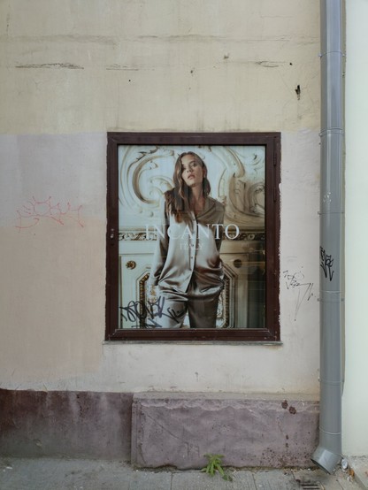 A  photo of an advertisement poster in a brown frame that has the text “INCANTO” and “ITALY” and features a model in a grey pantsuit type of clothing posing against a baroque background is on a white and yellowish wall of a building with a reddish bottom part. Next to the poster, there's a grey drainpipe.