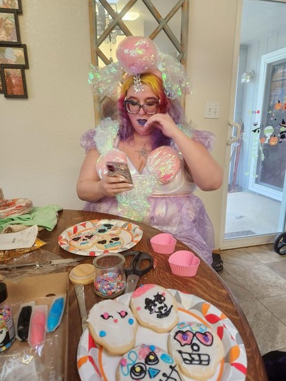 A picture of Stephanie looking down at her phone with her hand at her mouth in a costume behind a table full of Dia de Muertos skull cookies. She has long wavy yellow, pink, and purple hair with a big strip of jewels down her side part, matching eyebrows,  if glittery square rainbow leopard glasses, and purple lips. She is wearing a white body suit with big pink cellophane wrapped candies at the bust and on her head, and purple accents on the sleeves and skirt.