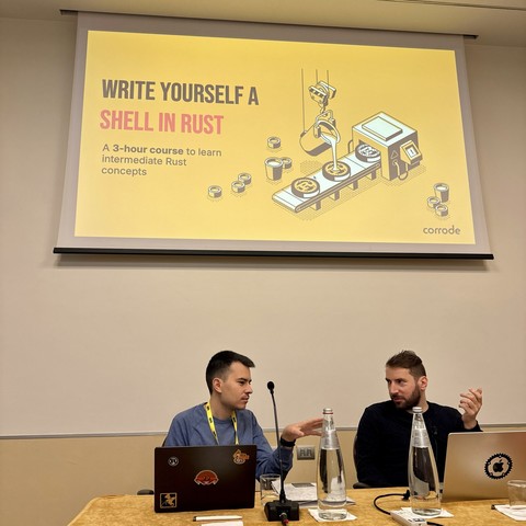 The image depicts two speakers seated at a table with laptops, engaged in discussion. A projector screen behind them displays the title "WRITE YOURSELF A SHELL IN RUST," promoting a 3-hour course on intermediate Rust concepts.