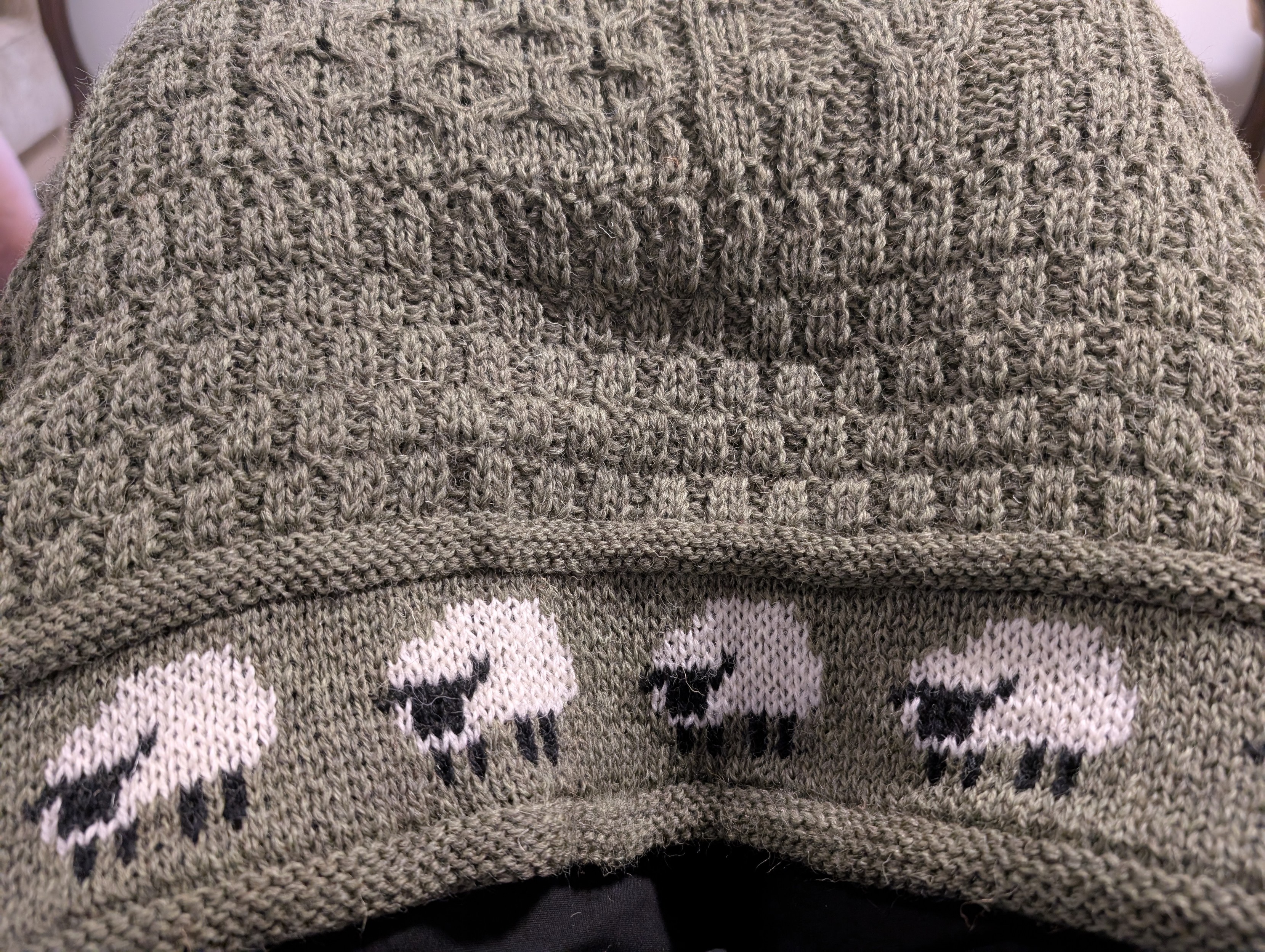 Photo of sage green throw, knitted in various patterns. Its border features a row of white sheep with black faces and legs.
