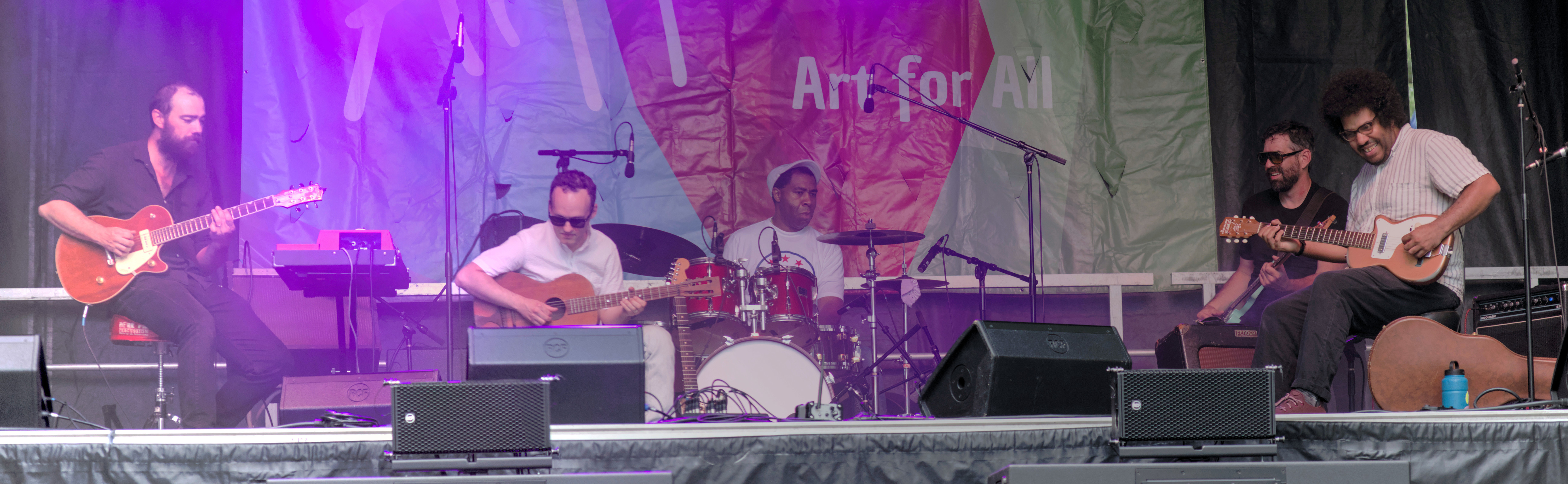 A panorama photo of a band playing on stage, composed of three guitar players, one drummer and one bass player, in various stages of enjoying their job.