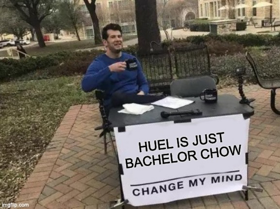 The image shows a man sitting at a table outdoors, wearing a blue long-sleeve shirt and holding a black mug. The table has a white banner with the text "HUEL IS JUST BACHELOR CHOW" and "CHANGE MY MIND" printed on it. There are two black mugs on the table, along with some papers and a black object that appears to be a microphone. The background features a park-like setting with trees, a building, and a paved walkway. The man is seated on a chair, and there is a bench behind him. The overall sett…