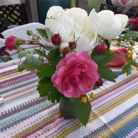 A bouquet of pink roses, white peonies, and small yellow wildflowers in a vase on a multicolored, striped tablecloth. 
