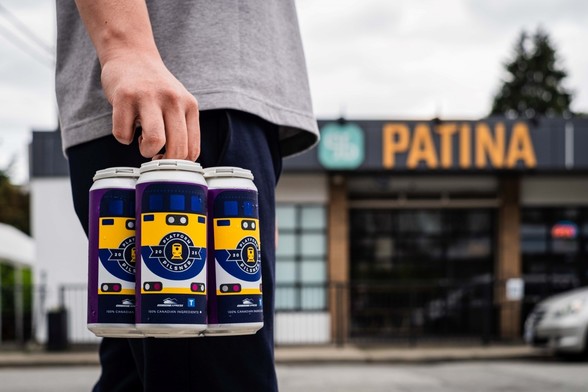 A man holding a four pack of beer cans in front of a building with the PATINA logo prominently displayed
