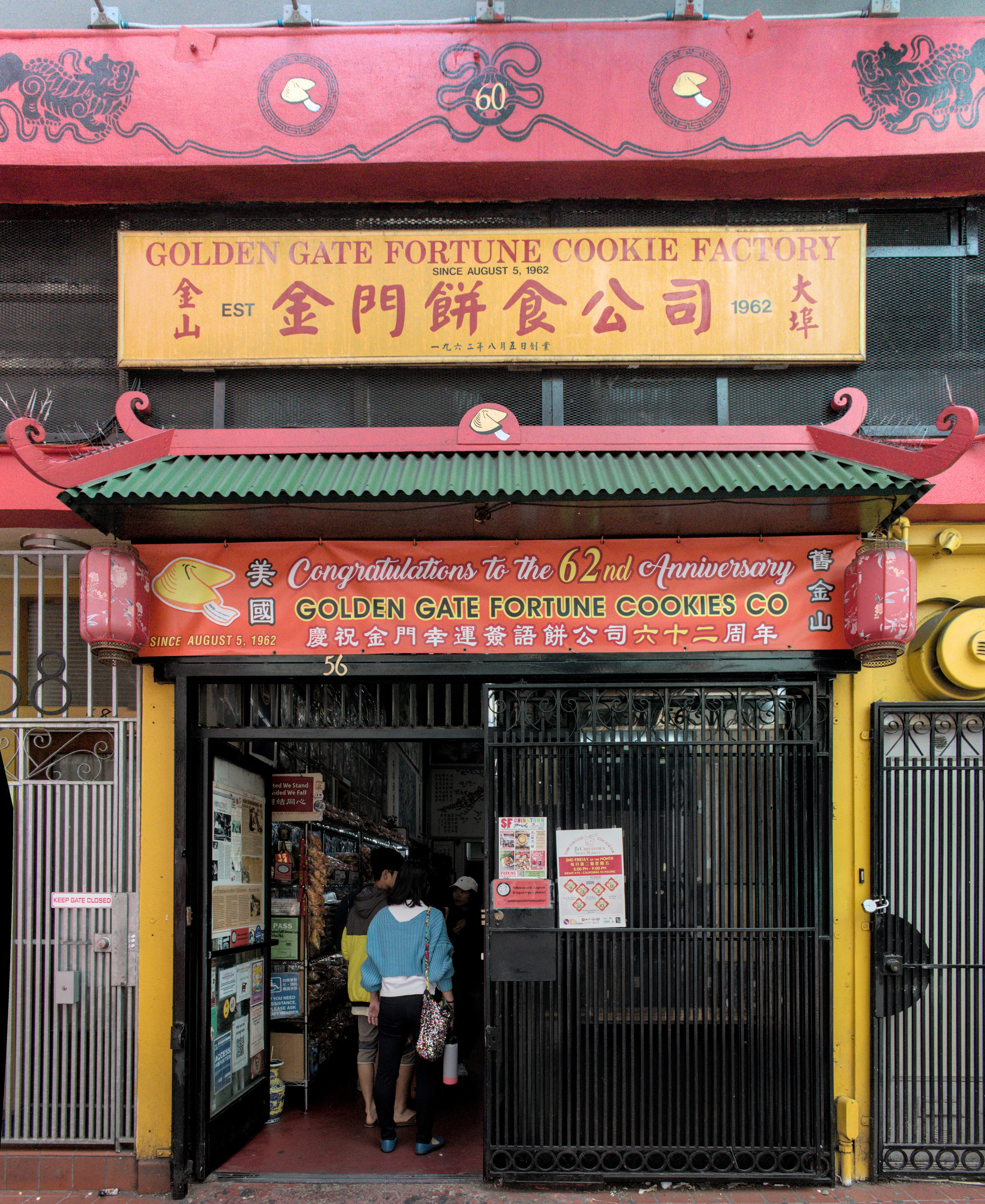 A photo of the front of a shop called GOLDEN GATE FORTUNE COOKIE FACTORY  A sign says Congratulations to the 62nd aniversary GOLDNE GATE FORTUNE COOKIE FACTORY, with a drawing of a fortune cookie  Some customers stand inside the doorway, looking at the products and at the workers making fortune cookies inside.