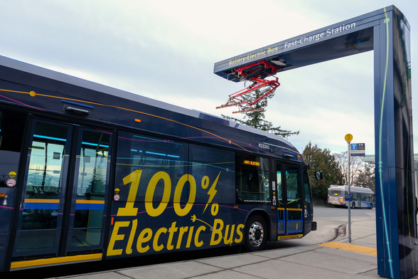 A new electric bus is parked underneath an electric charging device  which looks a lot like a pantograph. The side of the bus is decorated with a wrap which reads "100% Electric bus"