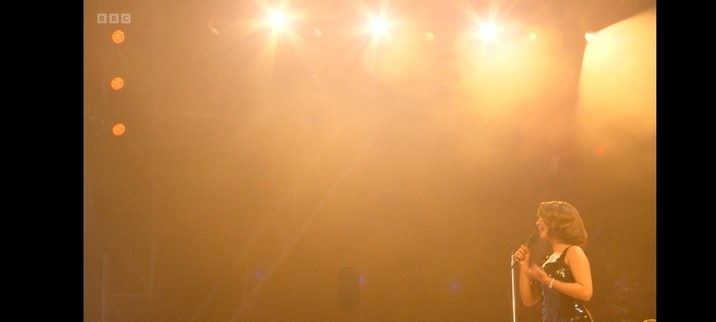 Raye in the bottom right corner of the frame, in a black sparkly dress, singing her heart out. The rest of the frame is washed with warm amber light.