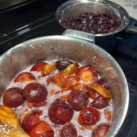 Two stainless steel pans of jam cooking on the stove. In the foreground are plums, in the background, cherries.
