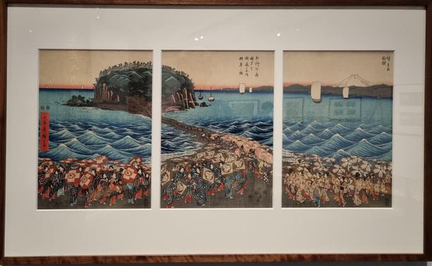 Three panel woodblock print, showing crowds in the foreground streaming towards a narrow isthmus crossing the emerald sea, towards a wooded island. There are many parasols.

Utagawa Hiroshige (1797-1858)
Crowds on Pilgrimage to See the Display
of Treasures at the Enoshima Benzaiten
Shrinein Sagami Province, 1851

弁
天
開
帳
登
阳
群
衆
図

Publishedby Sumiyoshiya Masagorö
Colour woodblockprint triptych
Giftfrom the collectionofAlanMedaugh
2025,AFBMLoan.1.5