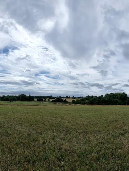 Wide clouded skies over a newly mown field, with a line between including trees, lake, and a stately home, just visible. The clouds are brightly sunlit, and show some dappled high cloud above.