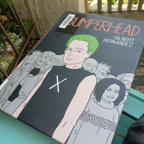 A hardback comic - Gilbert Hernandez, "Bumperhead" - sits on a turquoise chair with a porch and plants in the background. 
