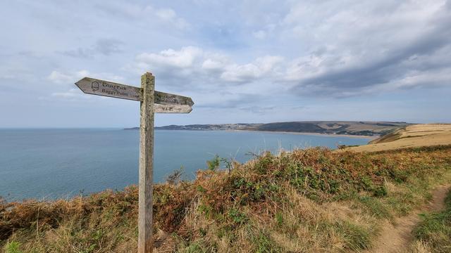 A wooden sign post on a walking path says 3 1/2 miles to Woolacombe, 1/2 mile to Baggy Point. Behind is a sweep of bay with teal blue sea. A town can be seen on the other side.
