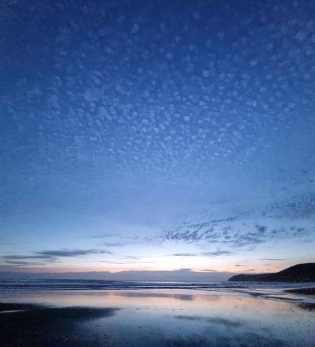 An evening shot of a vast, deep blue sky, dappled with tiny clouds that still catch the last of the sun's rays. Beneath lies a broad, water covered beach, reflecting the remaining dim sunset. A dark headland is to the right. It's all very moody and peaceful.