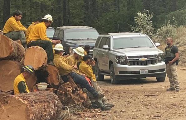 Bear Gulch wildfire fire crew/via Seattle Times
A group of men wearing white helmets and firefighter uniforms sit at the die of the road. A helmentless man stands facing them: he appears to be wearing a bullet proor vest and carrying a walkie talkie   