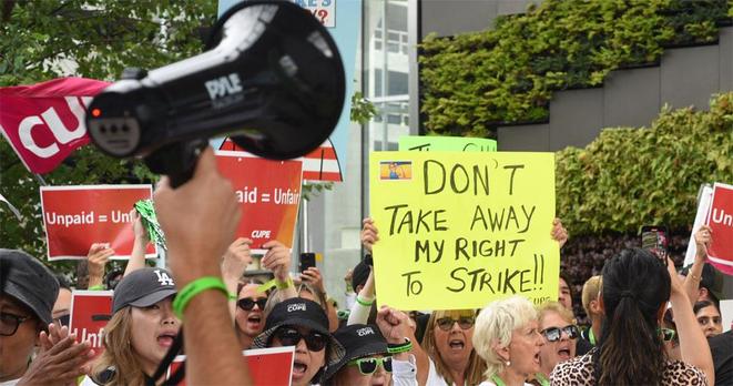 Women protesting outdoors holding signs that say "Unpaid = unfair" and prominently "DON'T TAKE AWAY MY RIGHT TO STRIKE" 

In the foreground a hand is holding up a megaphone  