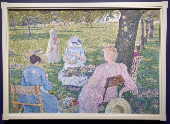 A group of women in an orchard, with the shadows falling straight down. They are busy with flowers and needlework, dressed in full summer dresses. One of the subjects is cut off behind a tree trunk.  The painting is pointillist, with dots of bright, contrasting colours that shimmer to the eye.
