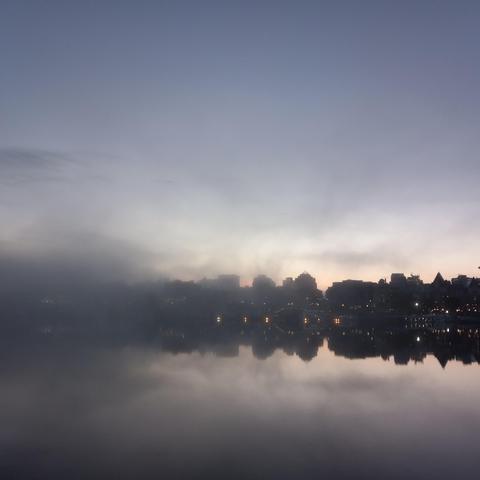 A city and its reflection on the still water obscured to the left by a thin veil of fog. Above is a pale blue sky showing the first pink and orange streaks of pre-dawn light.
