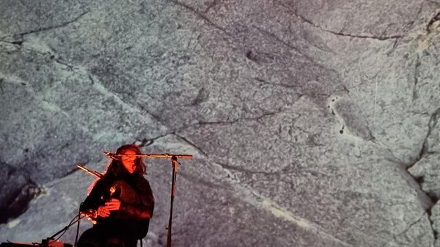 A woman in black plays the small-pipes in front of a massive projection of cracked grey stone. She is lit with a baleful red up-light, and her eyes are closed, head turned to the right, black hair over her eye.